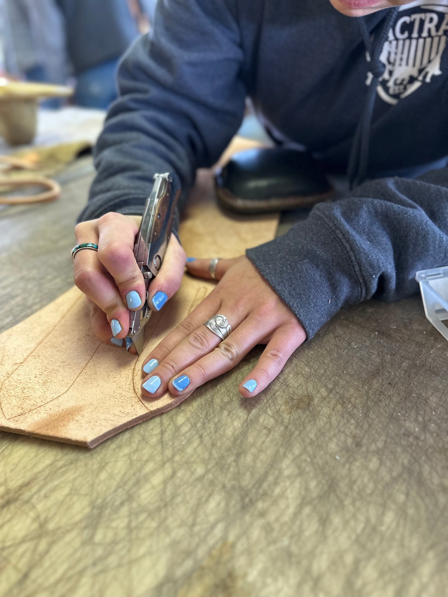 Person working on leather with a tool, wearing a dark hoodie and blue nail polish.