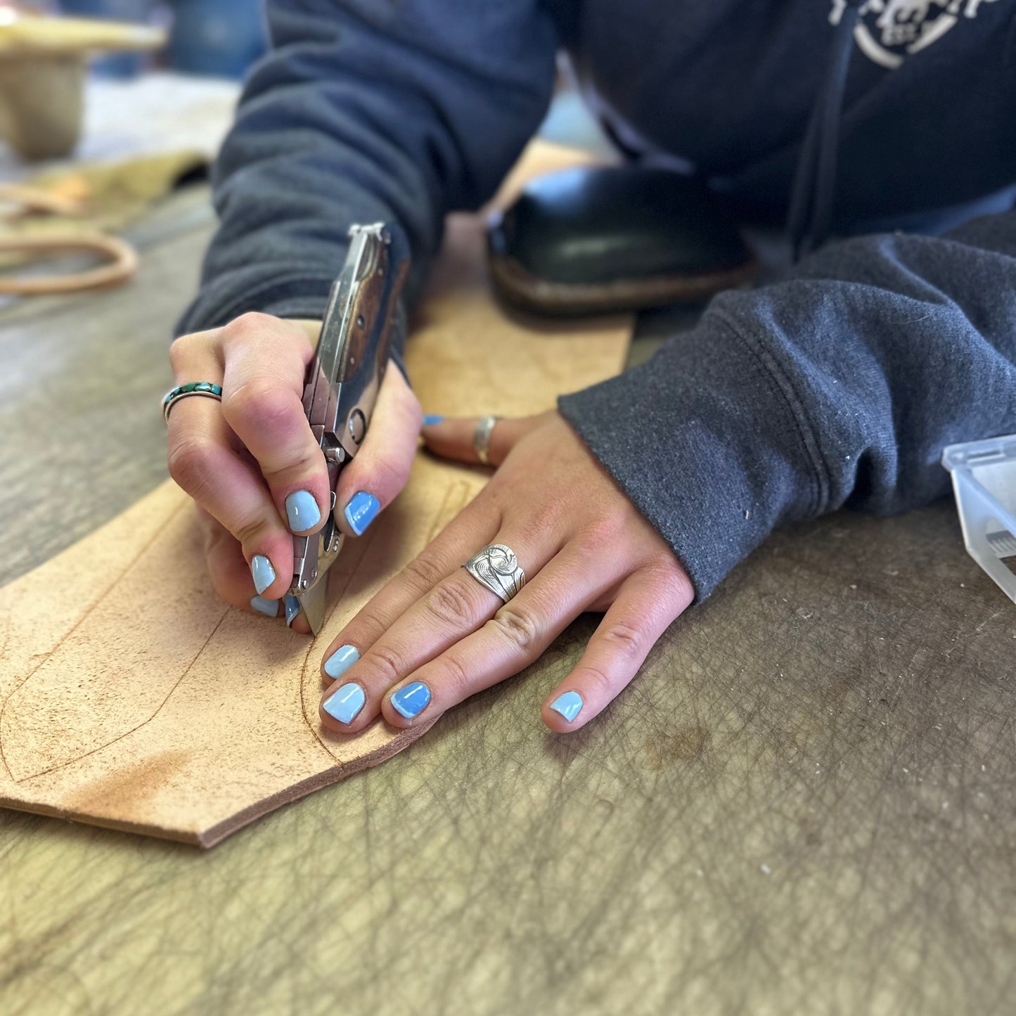 Person working on leather with a tool, wearing a dark hoodie and blue nail polish.