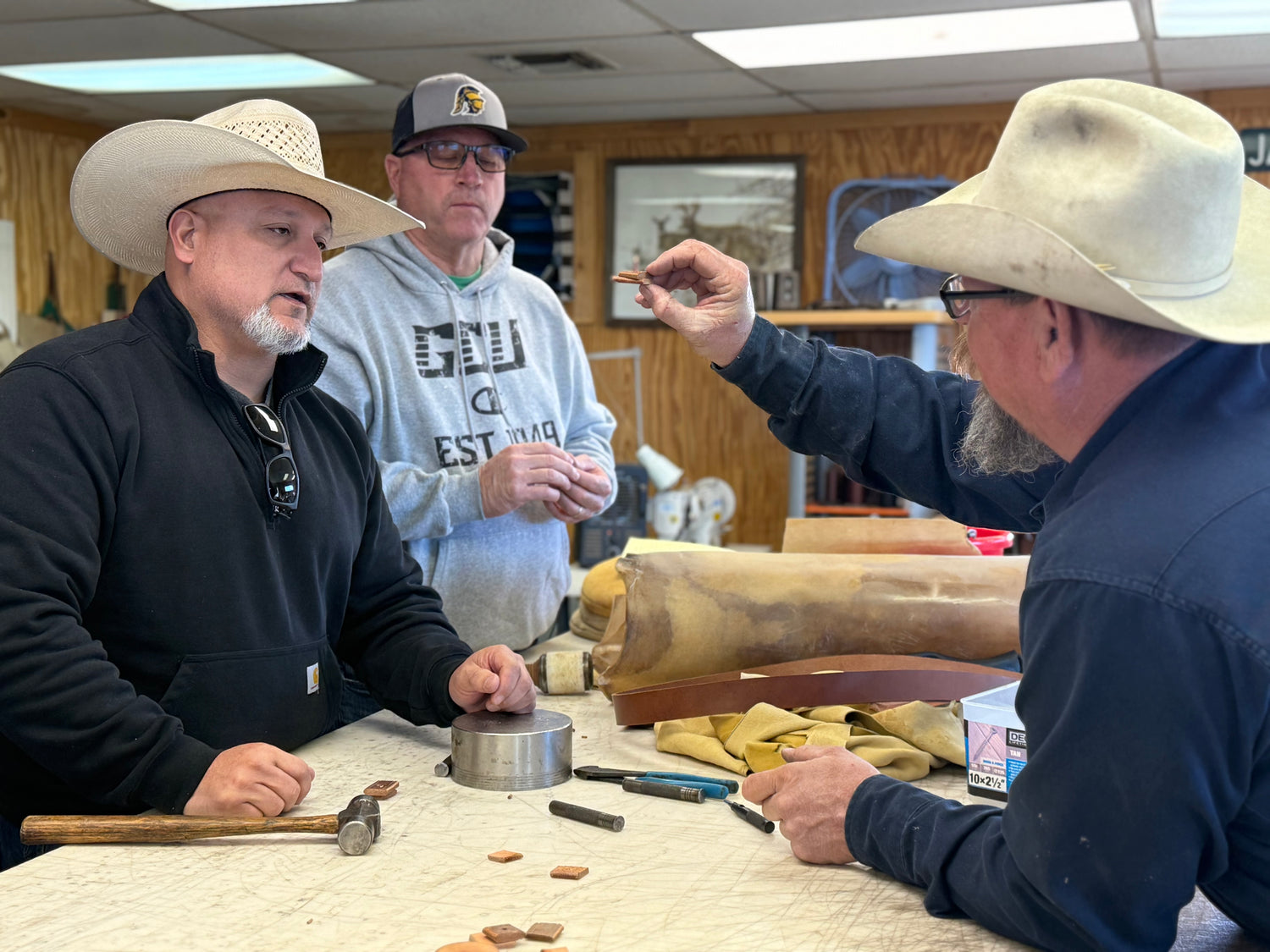 Three men in a workshop setting, with one using a hammer and another pointing at something on a table.