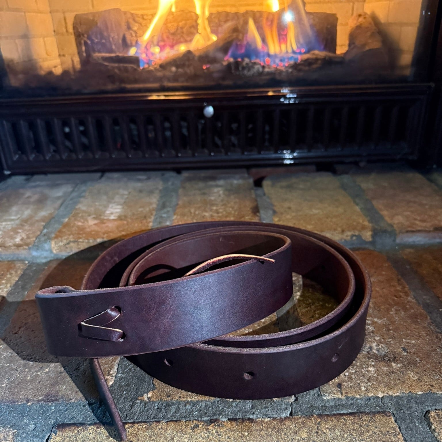 A brown Hermann Oak leather latigo belt is displayed in front of a fireplace. The belt is long and appears to be sturdy, with visible holes for sizing and a buckle for fastening.